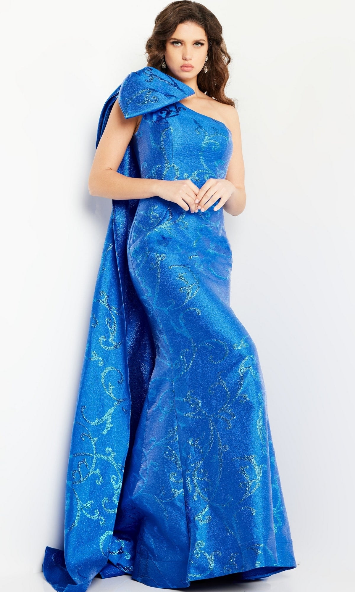 A woman poses against a white background in the Jovani One-Shoulder-Bow Long Formal Dress 23742, a bright blue, floor-length gown with a large bow detail and subtle floral patterns.
